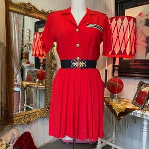 May include: A vintage red dress with short sleeves and a collared neckline. The dress features gold buttons, a black belt with a decorative buckle, and a pleated skirt. The dress is displayed on a mannequin in a vintage shop setting.