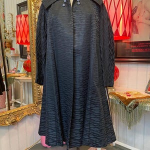 May include: A vintage black coat with a textured pattern, displayed on a mannequin. The coat features a wide collar and decorative buttons. The coat's design suggests a retro or mid-century style.