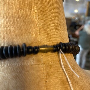 May include: Close-up of a black beaded necklace with a brass clasp. The necklace is displayed on a tan mannequin. White string hangs from the clasp.