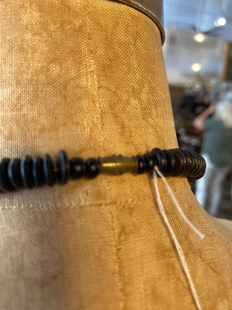 May include: Close-up of a black beaded necklace with a brass clasp. The necklace is displayed on a tan mannequin. White string hangs from the clasp.