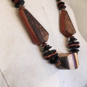 May include: A wooden necklace featuring geometric shapes and disc-shaped beads in shades of brown, black, and tan. The necklace has a black cord and is displayed on a mannequin.