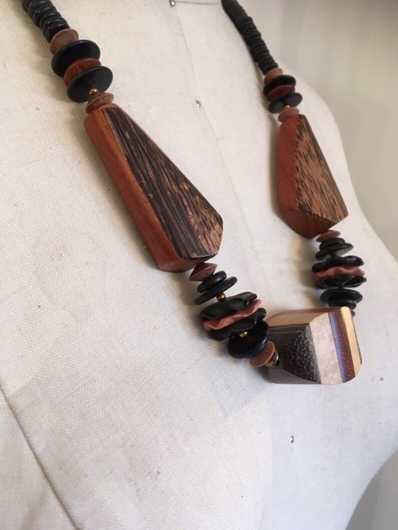 May include: A wooden necklace featuring geometric shapes and disc-shaped beads in shades of brown, black, and tan. The necklace has a black cord and is displayed on a mannequin.