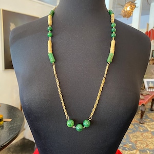 May include: A gold chain necklace with green and beige beads. The necklace is displayed on a black mannequin.