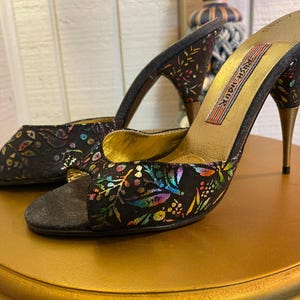 May include: A pair of black high-heeled mules with a floral pattern in various colors. The heels are gold-colored, and the insoles are gold. The text "Rush Hour" is printed on the insole. The shoes are on a gold-colored surface.