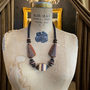 May include: A wooden necklace with geometric shapes and a mix of dark brown, light brown, and black beads. The necklace is displayed on a vintage mannequin. The necklace features a central oval pendant with striped details.