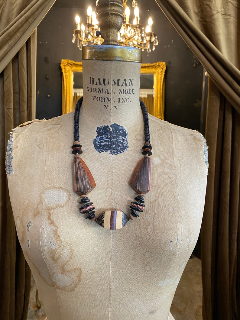 May include: A wooden necklace with geometric shapes and a mix of dark brown, light brown, and black beads. The necklace is displayed on a vintage mannequin. The necklace features a central oval pendant with striped details.