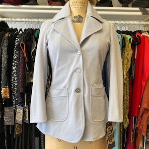 May include: A light blue blazer with a notched collar, two front pockets, and three buttons. The jacket is displayed on a vintage mannequin in a clothing store, with other garments visible in the background.