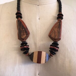 May include: A wooden necklace with a variety of shapes and colors. The necklace features large, geometric wooden pendants in shades of brown and black, connected by a strand of black beads. The central pendant is oval-shaped with vertical stripes.