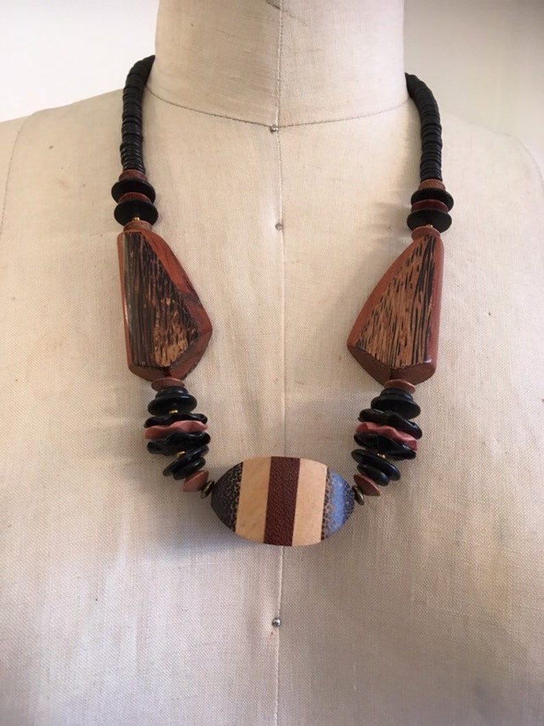May include: A wooden necklace with a variety of shapes and colors. The necklace features large, geometric wooden pendants in shades of brown and black, connected by a strand of black beads. The central pendant is oval-shaped with vertical stripes.