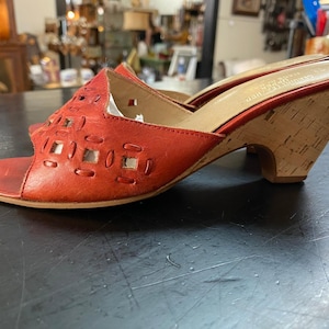 May include: A pair of red leather slide sandals featuring a cutout geometric pattern on the upper strap, an open toe design, and a light-colored cork-textured wedge heel. The interior footbed is tan and includes visible text branding.