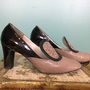 K&ouml;nnte beinhalten: Ein Paar braune und schwarze Schuhe mit Absatz und einem dekorativen Detail an der Spitze. Die Schuhe haben ein schwarzes Lackleder-Obermaterial und ein braunes Leder-Untermaterial.