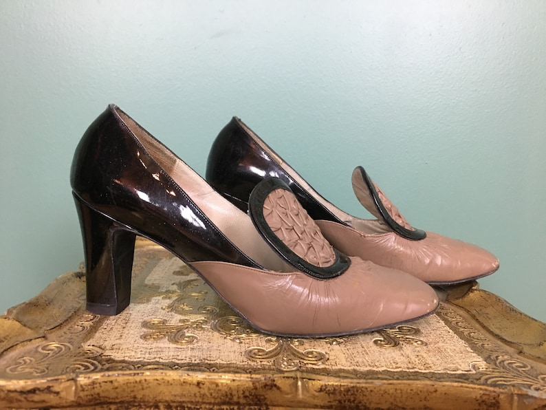 K&ouml;nnte beinhalten: Ein Paar braune und schwarze Schuhe mit Absatz und einem dekorativen Detail an der Spitze. Die Schuhe haben ein schwarzes Lackleder-Obermaterial und ein braunes Leder-Untermaterial.
