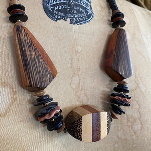 May include: A wooden necklace with a variety of shapes and colors. The necklace features large, geometric wooden pendants in shades of brown and black, interspersed with smaller, round beads. The necklace is displayed on a mannequin.