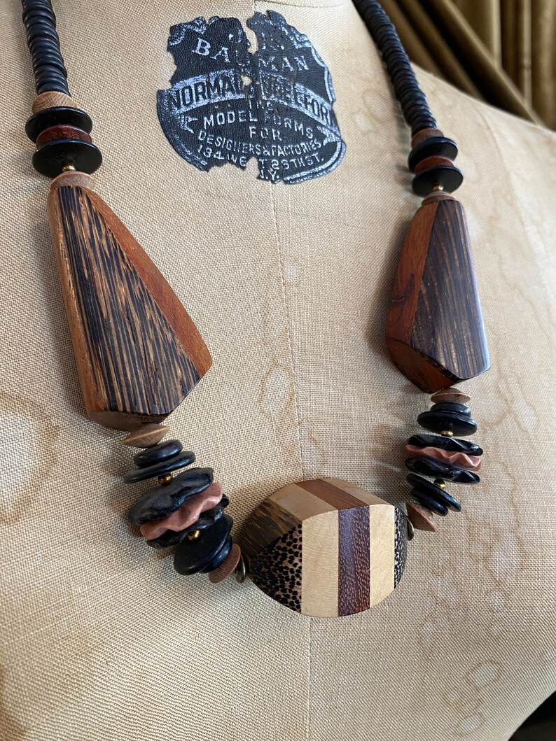 May include: A wooden necklace with a variety of shapes and colors. The necklace features large, geometric wooden pendants in shades of brown and black, interspersed with smaller, round beads. The necklace is displayed on a mannequin.