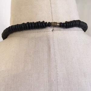May include: A black beaded necklace with a brass clasp, displayed on a mannequin. The necklace is composed of numerous small, round, dark beads. The clasp is a contrasting metallic color. The necklace is a fashion accessory.