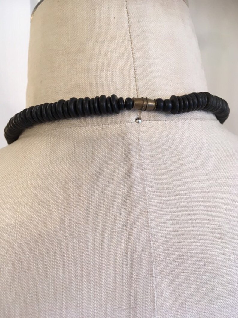May include: A black beaded necklace with a brass clasp, displayed on a mannequin. The necklace is composed of numerous small, round, dark beads. The clasp is a contrasting metallic color. The necklace is a fashion accessory.