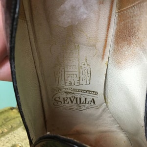 K&ouml;nnte beinhalten: Nahaufnahme des Innenraums eines schwarzen Schuhs mit einer braunen Ledersohle. Die Innensohle tr&auml;gt ein goldenes Logo mit der Aufschrift "SEVILLA" und einer Zeichnung eines Geb&auml;udes und einer Br&uuml;cke.