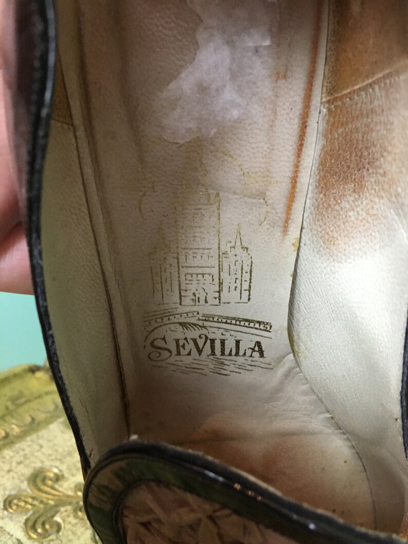 K&ouml;nnte beinhalten: Nahaufnahme des Innenraums eines schwarzen Schuhs mit einer braunen Ledersohle. Die Innensohle tr&auml;gt ein goldenes Logo mit der Aufschrift "SEVILLA" und einer Zeichnung eines Geb&auml;udes und einer Br&uuml;cke.