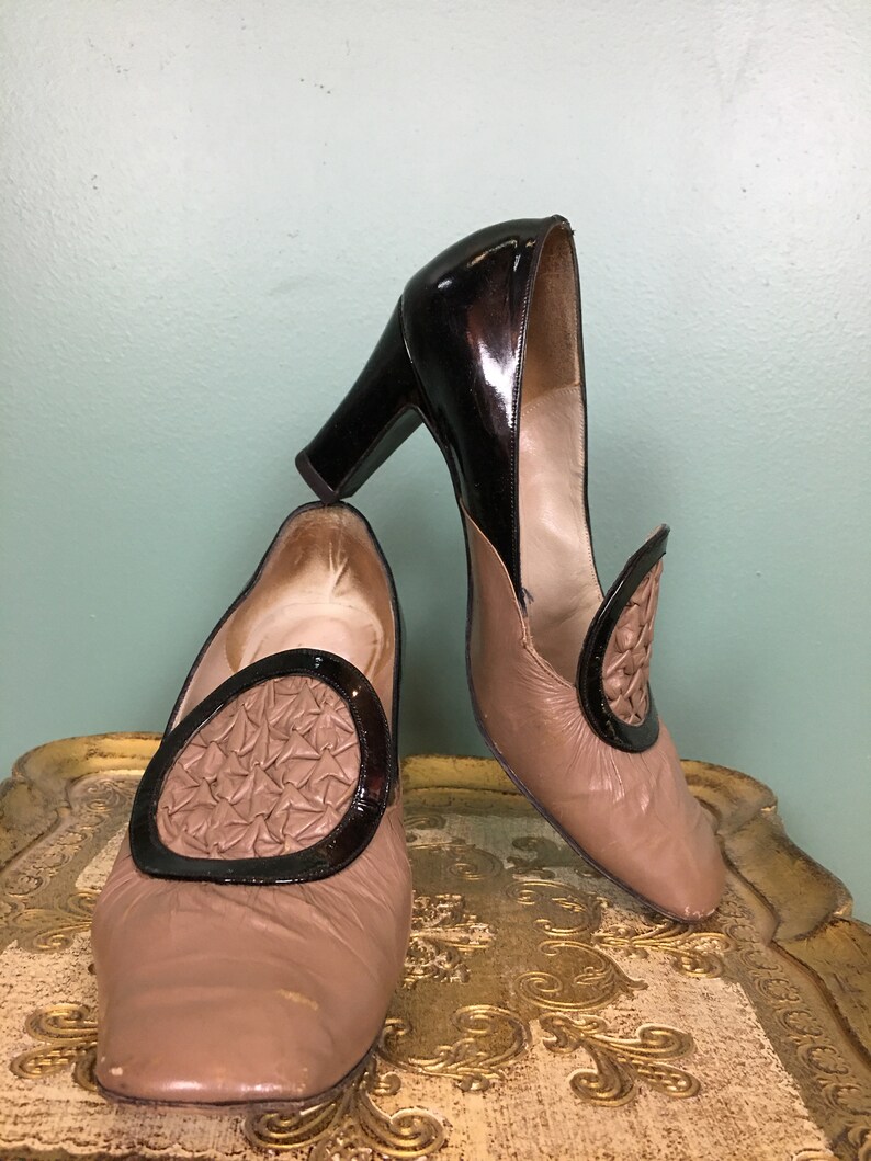 K&ouml;nnte beinhalten: Ein Paar braune Lederschuhe mit schwarzen Lackleder-Akzenten. Die Schuhe haben eine quadratische Spitze und ein dekoratives Design auf der Vorderseite.