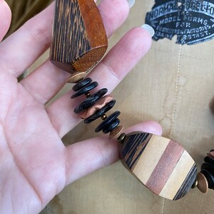 May include: A handcrafted wooden necklace featuring geometric shapes and a mix of colors. The necklace includes dark brown, light brown, and copper-toned beads and pendants. The design showcases a unique, artistic style.