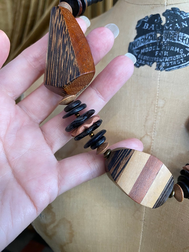 May include: A handcrafted wooden necklace featuring geometric shapes and a mix of colors. The necklace includes dark brown, light brown, and copper-toned beads and pendants. The design showcases a unique, artistic style.