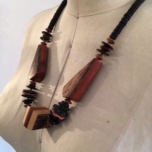 May include: A wooden necklace with geometric and organic shapes. The necklace features large, reddish-brown wooden pendants and black, brown, and pink beads. The necklace is displayed on a mannequin.