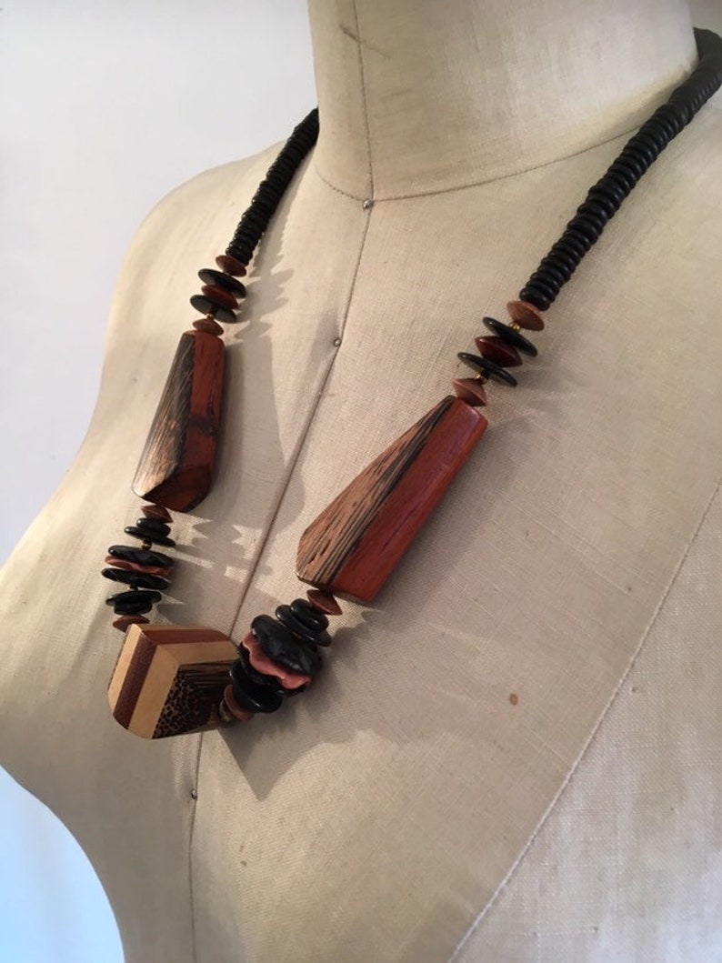 May include: A wooden necklace with geometric and organic shapes. The necklace features large, reddish-brown wooden pendants and black, brown, and pink beads. The necklace is displayed on a mannequin.