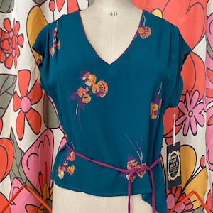 Puede incluir: Blusa vintage de color verde azulado con cuello en V y mangas cortas. La blusa presenta un estampado floral en tonos naranja, amarillo y morado. Un cinturón morado a juego ciñe la cintura. Una etiqueta con el texto "BLACK LABEL VINTAGE" cuelga de la blusa.