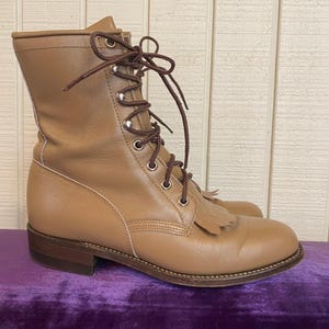 May include: A tan leather lace-up boot featuring a mid-calf height, a rounded toe, and a decorative fringe panel over the laces. The boot includes dark brown laces threaded through silver-toned metal eyelets and a stacked brown heel.