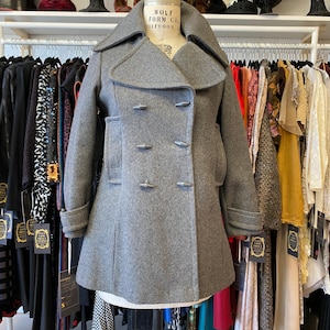 May include: A gray double-breasted peacoat with a wide collar and a flared hem. The coat is on a mannequin in a clothing store.