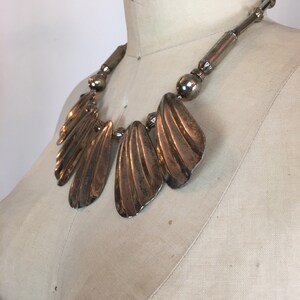 May include: A silver statement necklace with a large, fan-shaped pendant. The pendant is made of multiple overlapping pieces of metal, creating a textured and organic look.