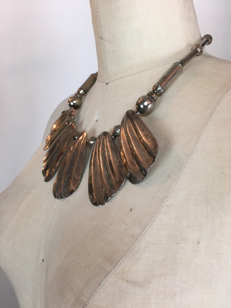 May include: A silver statement necklace with a large, fan-shaped pendant. The pendant is made of multiple overlapping pieces of metal, creating a textured and organic look.