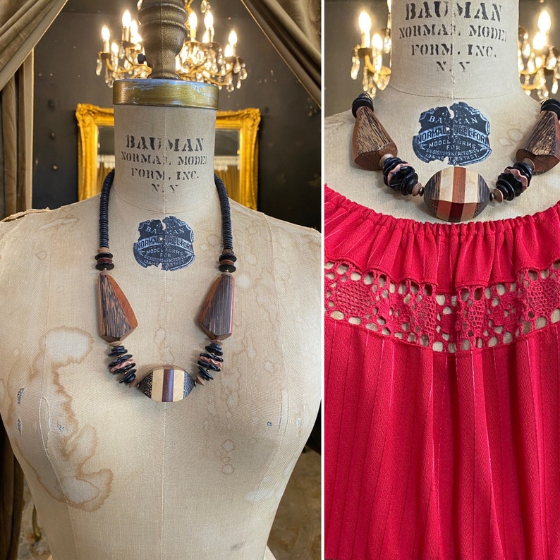 May include: A statement necklace with large wooden geometric shapes and beads in shades of brown, black, and tan. The necklace is displayed on a vintage dress form. The dress form has the text "BAUMAN NORMAN MODE FORM. INC. N.Y." printed on it.