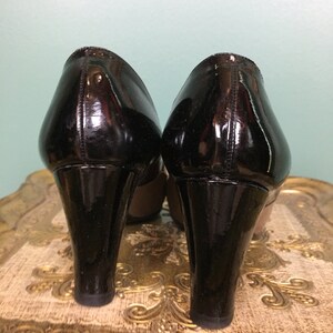 K&ouml;nnte beinhalten: Ein Paar schwarze Lackleder-Plateau-Heels mit hohem Absatz. Die Abs&auml;tze befinden sich auf einer goldfarbenen, gemusterten Oberfl&auml;che.