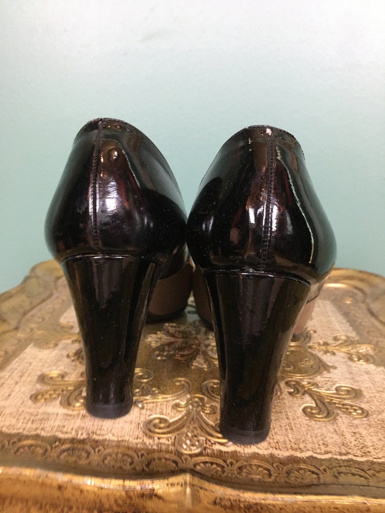 K&ouml;nnte beinhalten: Ein Paar schwarze Lackleder-Plateau-Heels mit hohem Absatz. Die Abs&auml;tze befinden sich auf einer goldfarbenen, gemusterten Oberfl&auml;che.