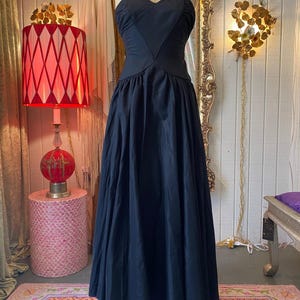 May include: A floor-length black gown featuring a fitted bodice with a sweetheart neckline, thin spaghetti straps, and a full, pleated skirt. The dress is displayed on a mannequin and includes a dark beaded necklace at the neckline.