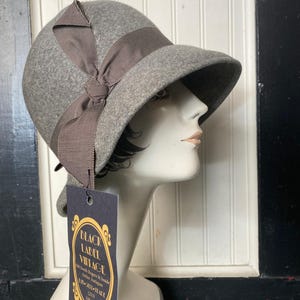 May include: A grey felt cloche hat featuring a brown grosgrain ribbon band and a side bow. A dark rectangular tag hangs from the hat, printed with the text: BLACK LABEL VINTAGE, 308 South Rogers St Inside, Atelier Antiques, BUY-SELL-TRADE, (253) 348-5252.