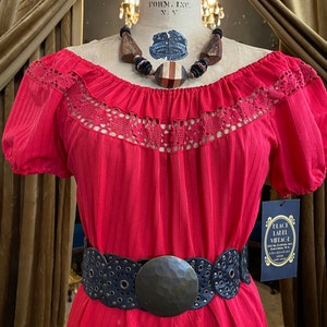 May include: A vintage red dress with a crochet neckline and short sleeves, cinched at the waist with a black belt featuring a large, textured buckle. A wooden beaded necklace and a Black Label Vintage tag complete the look.