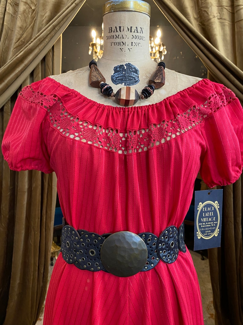 May include: A vintage red dress with a crochet neckline and short sleeves, cinched at the waist with a black belt featuring a large, textured buckle. A wooden beaded necklace and a Black Label Vintage tag complete the look.