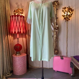 May include: A light mint green knee-length robe featuring short sleeves with beige lace trim at the cuffs and a matching beige lace collar. The garment has a front button-down closure with five small buttons extending from the neckline to the waist.