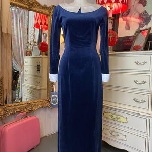 May include: A navy blue velvet dress with a cream-colored collar and cuffs. The long-sleeved dress is displayed on a mannequin. The dress has a classic, elegant design, suitable for formal occasions. The dress is floor-length.