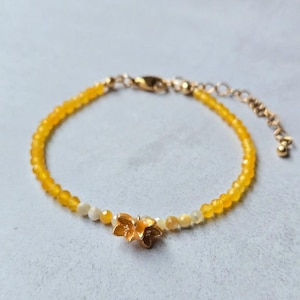 Puede incluir: Una pulsera delicada con pequeñas cuentas facetadas amarillas y blancas translúcidas dispuestas en un patrón circular. Un dije de flor de metal dorado está centrado en la hebra, que incluye una cadena de metal dorado con cierre de mosquetón.