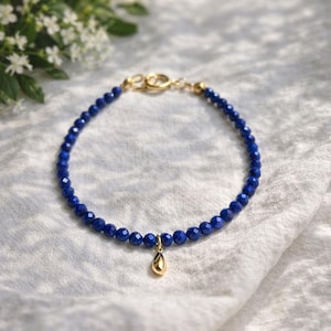 Pode incluir: Uma pulseira delicada com pequenas contas facetadas azul-escuras. A pulseira tem um fecho dourado e inclui um pequeno pingente dourado em forma de gota. A joia é exibida em uma superfície branca texturizada.
