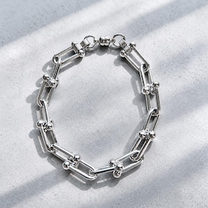 May include: A silver bracelet with a chain link design. The bracelet features a series of rectangular links connected by smaller, rounded links. The links are adorned with small, spherical accents. The bracelet is displayed on a light gray surface.