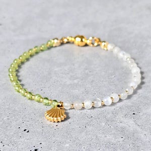 May include: A beaded bracelet with a gold seashell charm. The bracelet features light green and white beads, with gold accents. The clasp is also gold. The bracelet is displayed on a grey surface.