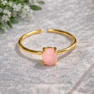 Pode incluir: Um anel ajustável dourado com uma pedra preciosa oval rosa claro. A banda do anel é aberta no topo, e a pedra preciosa é mantida no lugar por quatro garras. O anel está sobre um fundo macio e texturizado.