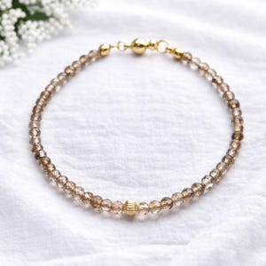 Puede incluir: Una delicada pulsera con pequeñas cuentas facetadas de cuarzo ahumado. La pulsera tiene un cierre magnético de color dorado y una sola cuenta dorada más grande con un diseño texturizado. La pulsera se muestra sobre una superficie blanca.
