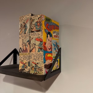 May include: A vintage Superman comic book with a yellow, red, and blue cover featuring the title Giant Superman and the text Stop! You Must See These Secret Pages. The comic is displayed upright on a black metal shelf bracket against a wall.