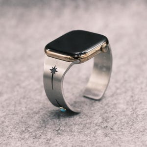 Könnte beinhalten: Eine Smartwatch mit goldfarbenem Metallgehäuse und schwarzem Display, befestigt an einem Armreif aus gebürstetem silberfarbenem Metall. Das Armband zeigt ein eingraviertes schwarzes Sternenmotiv und einen kleinen, runden Türkis-Stein.
