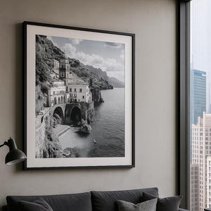 May include: A framed black and white photograph of a coastal town with buildings on a cliffside, a beach, and the sea. The photograph hangs above a dark gray sofa with throw pillows and a round coffee table. A window shows a city skyline.